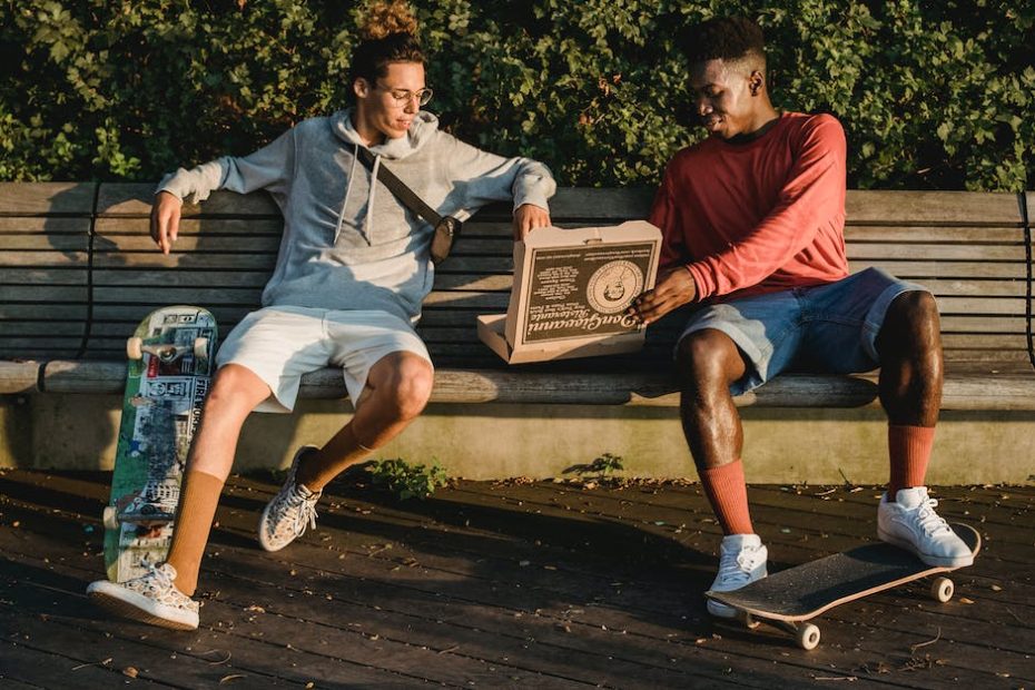 Full body content multiracial male friends with skateboards wearing casual shorts sitting on wooden bench and enjoying tasty fresh pizza in park