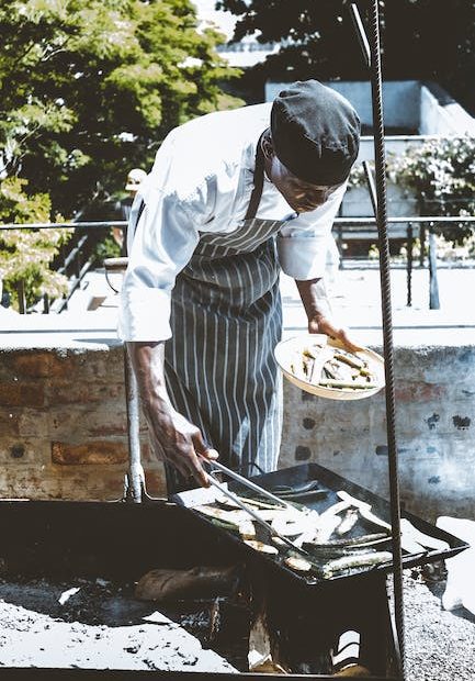 African American male cook in apron standing near railing and grilling food in sunny summer day