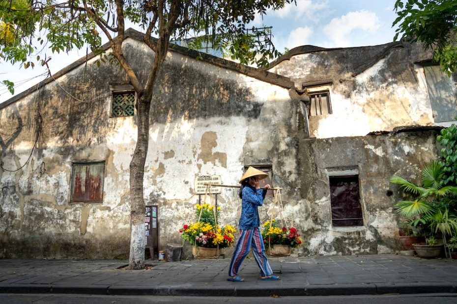 Asian woman walking near shabby buildings and carrying baskets with flowers on street