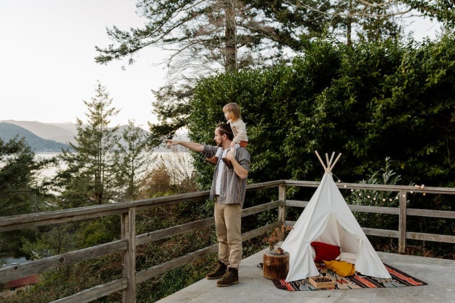 Side view of little child sitting on shoulders of father while man pointing to mountain while standing on wooden terrace with wigwam and picturesque view