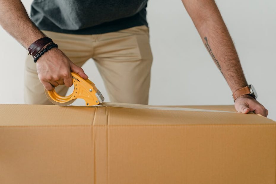 Crop anonymous young male packer in casual clothes using tape gun dispenser for sealing cardboard boxes