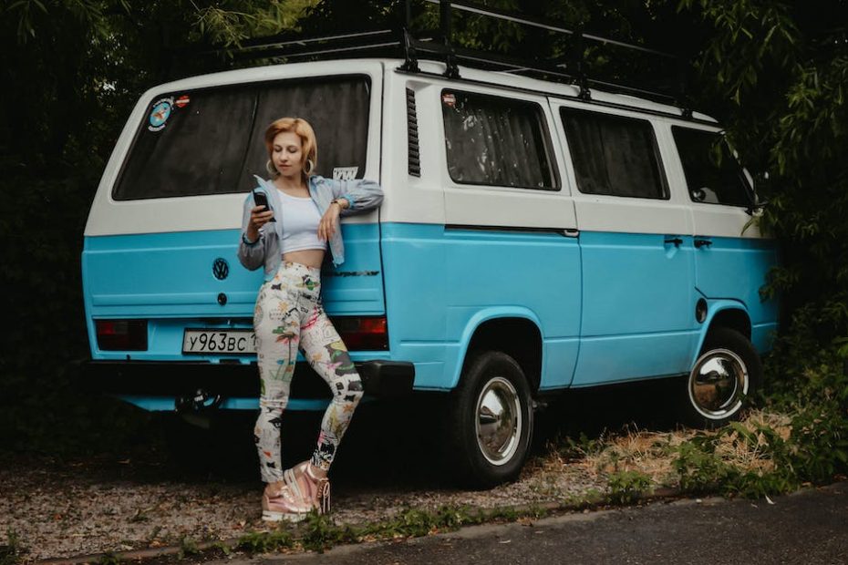 Full length of young female in vintage outfit dialing number on wireless handset while standing near minivan