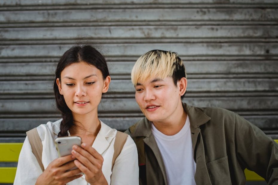 Content young Asian couple in casual outfits surfing contemporary mobile phones and sitting on street bench on clear weather