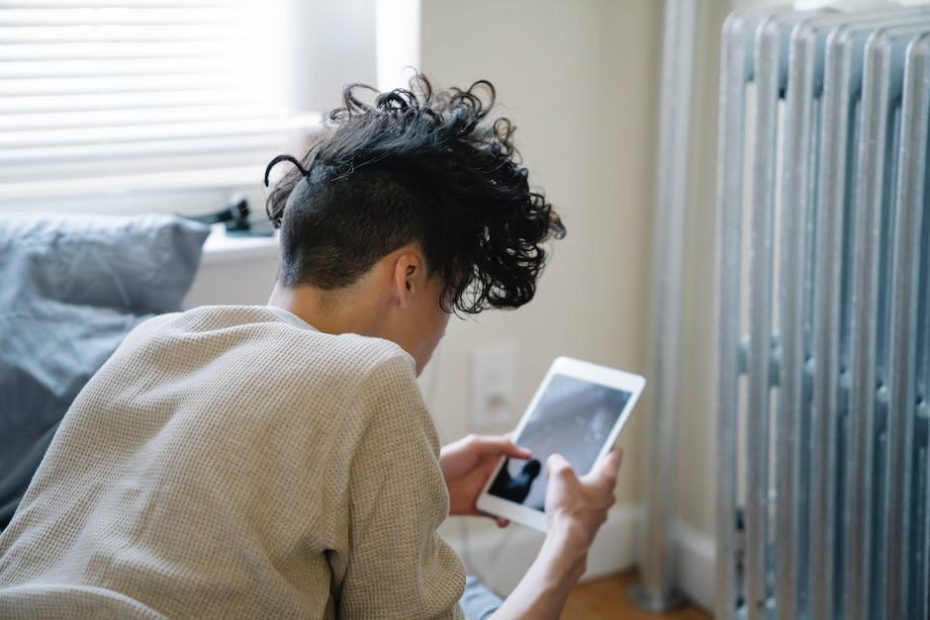 Unrecognizable young man with curly hair lying on bed and texting on tablet in weekend morning at home